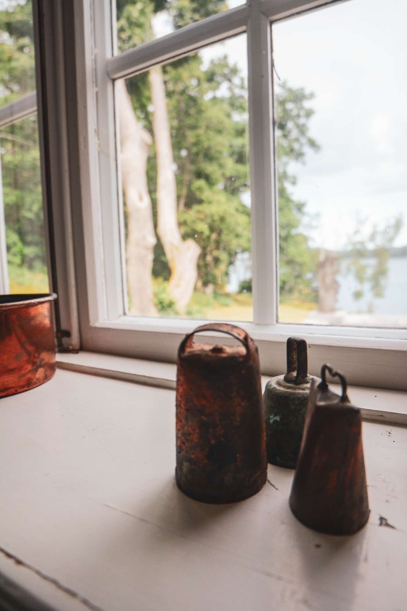 Tre gamla koskällor i järn och koppar på en vit fönsterbänk, med utsikt genom fönstret mot trädstammar och vattnet.