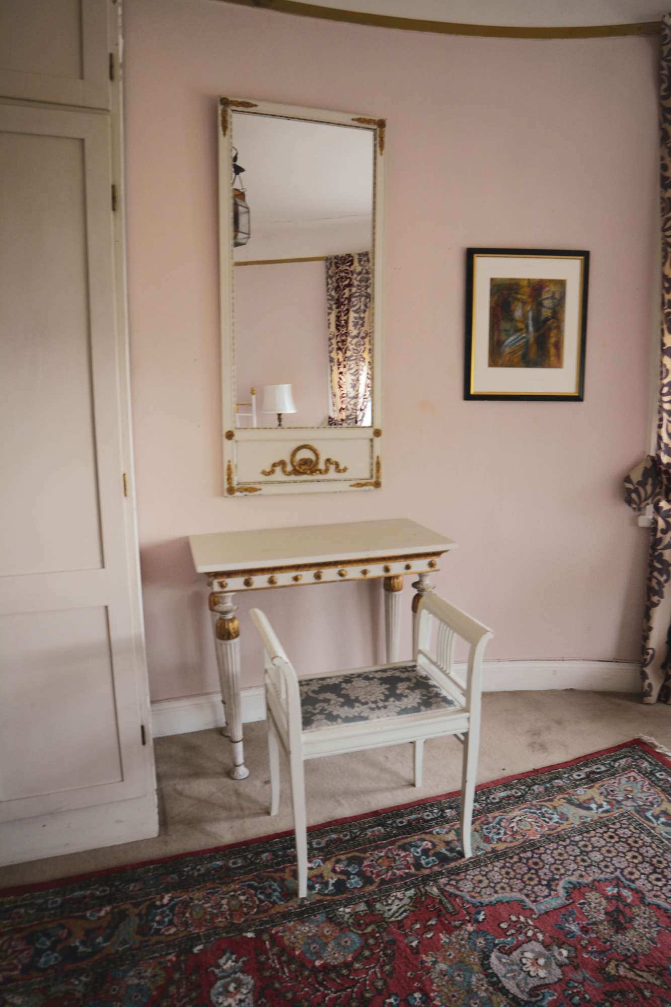 Rosa vägg med en hög förgylld spegel över ett vitt toalettbord och en stol, en målning bredvid och en röd orientalisk matta på golvet.