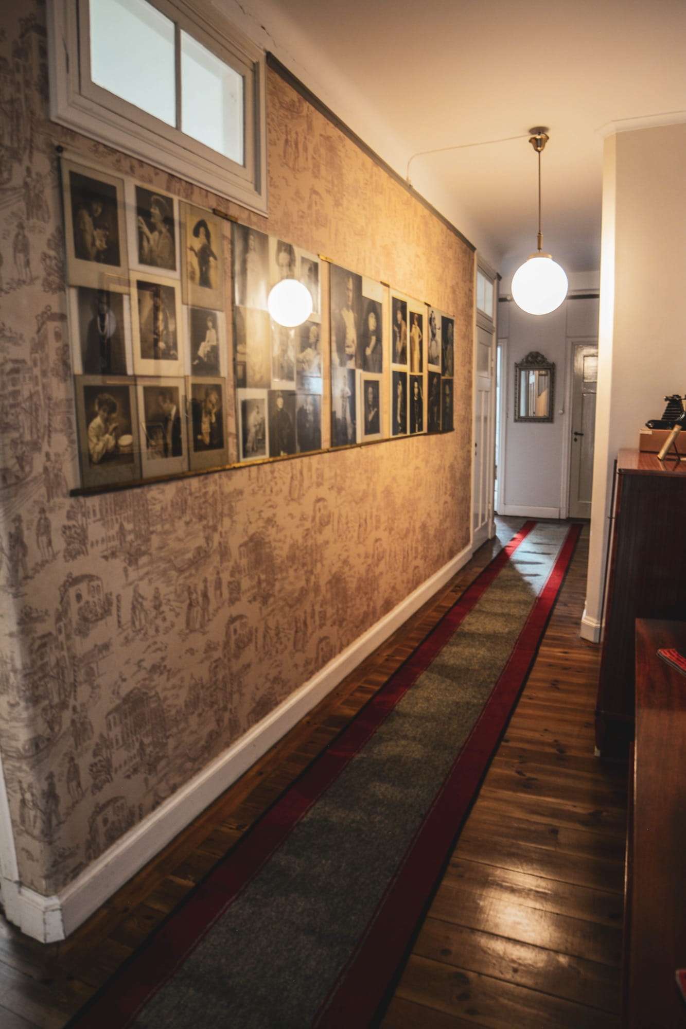 Korridor med figurtapet och en hel vägg av inramade svartvita porträttfotografier, glasklot i taket och en löpare på plankgolvet.