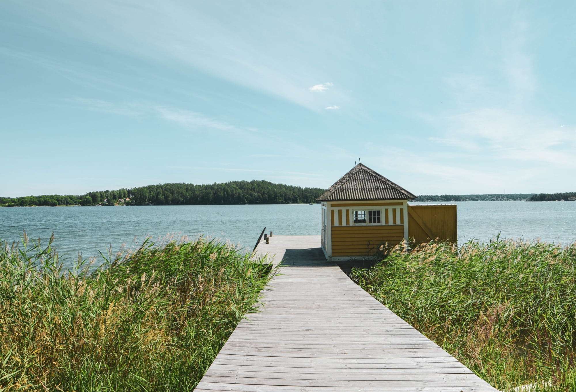 Bryggan rakt ut genom vassen till det gula badhuset, med öppen sjö och öar bortom.