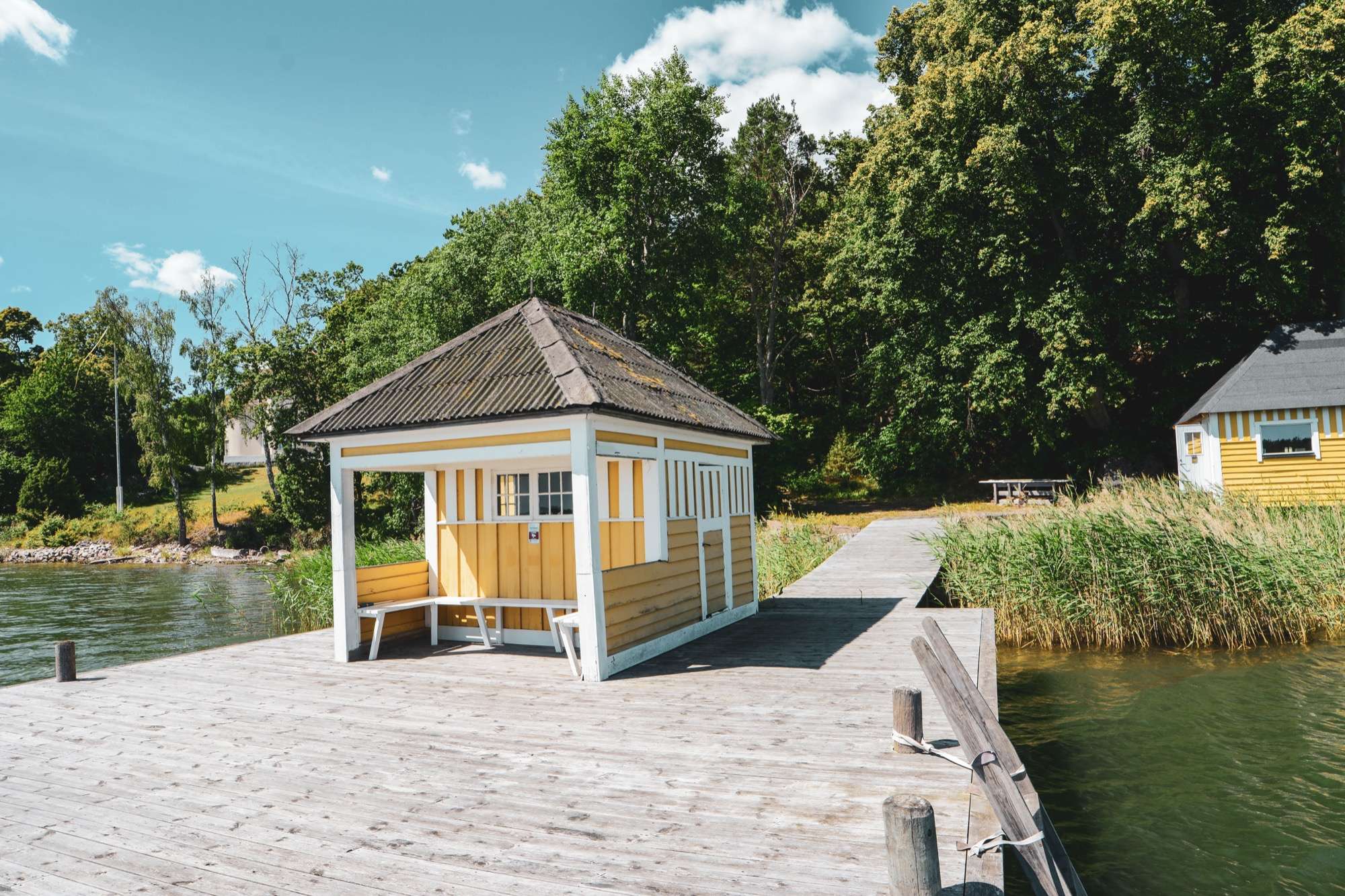 Bryggan i trä med en gul paviljong med vitmålade knutar och bänkar, vass längs kanten och badhuset i gult längre bort.