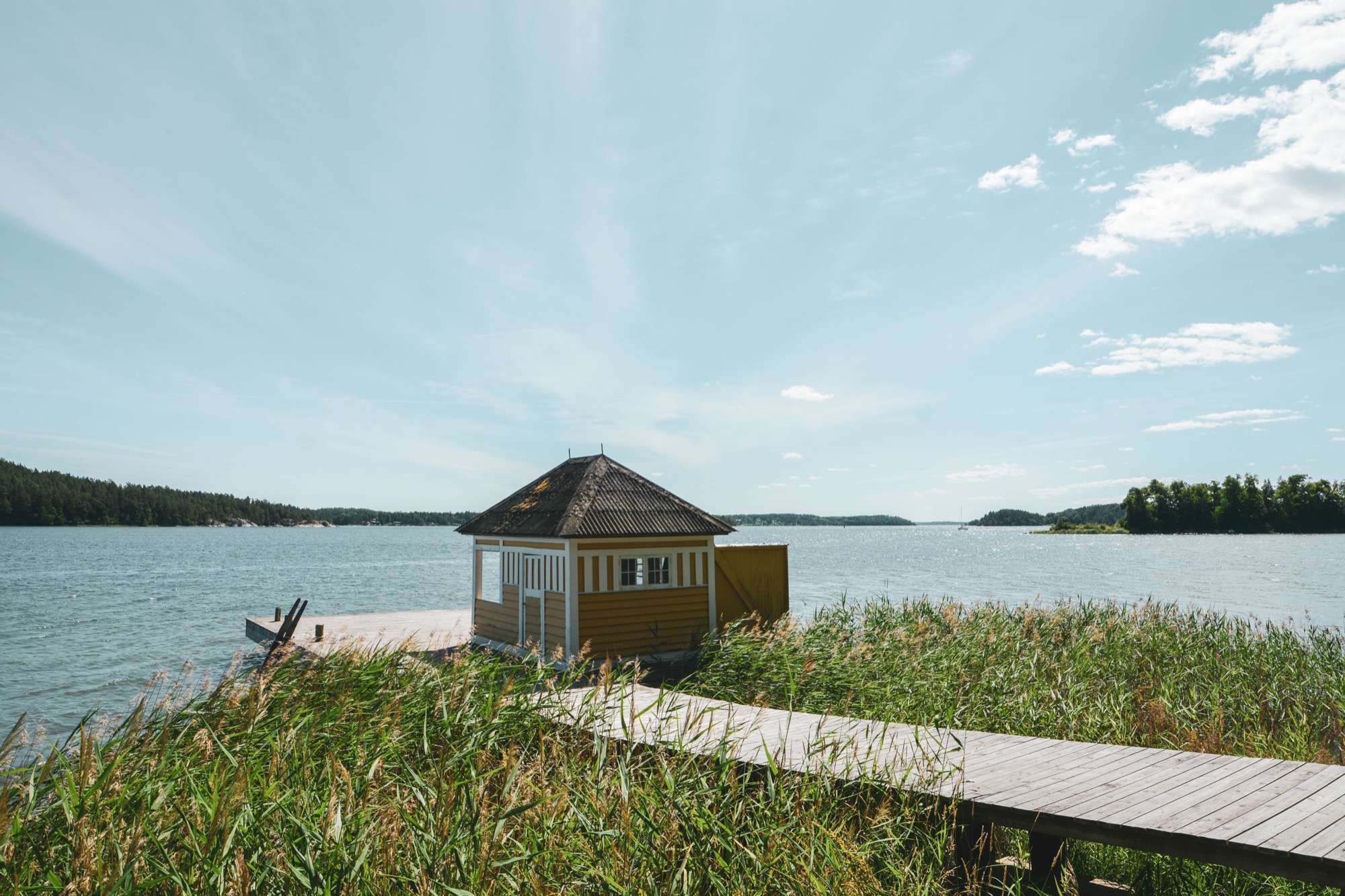 Det gula badhuset längst ut på bryggan, med vasskanten i förgrunden och öppet vatten och skogsklädda öar bortom.
