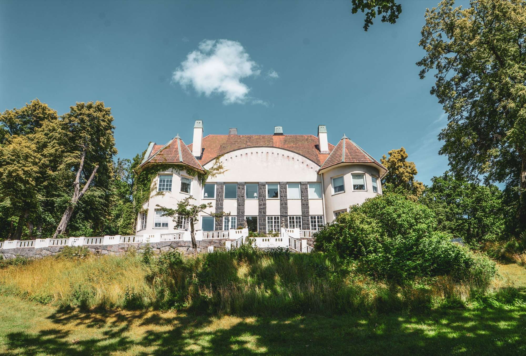 Herrhaga sett från gräset på sjösidan. Ljus putsad fasad, rött valmat tak, två runda hörntorn och en vit balustrad längs terrassen, under blå himmel.