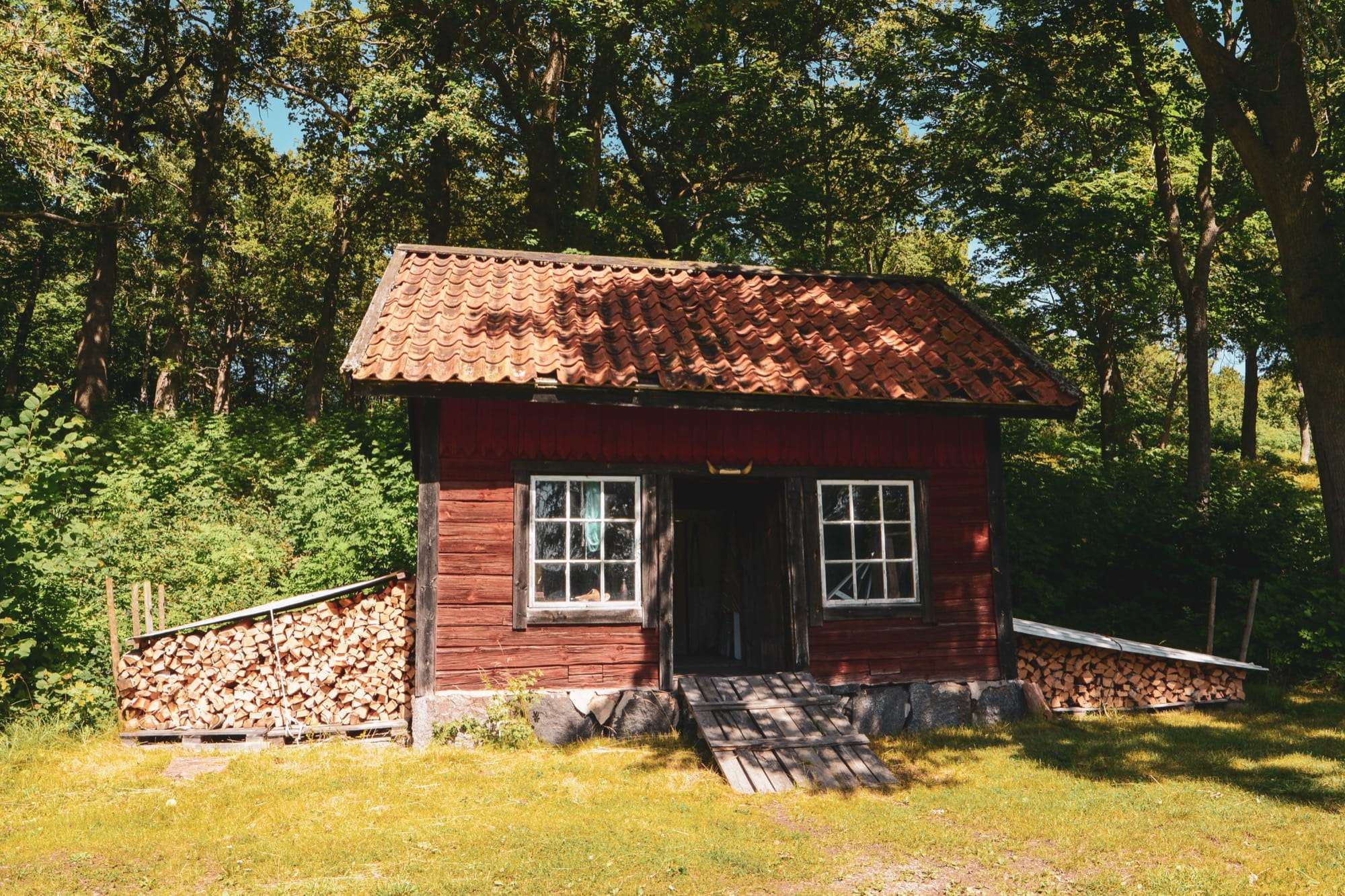 Liten röd timmerstuga med tegeltak och vita spröjsade fönster i skogskanten, med ved staplad under tak på båda sidor och en trätrall framför dörren.