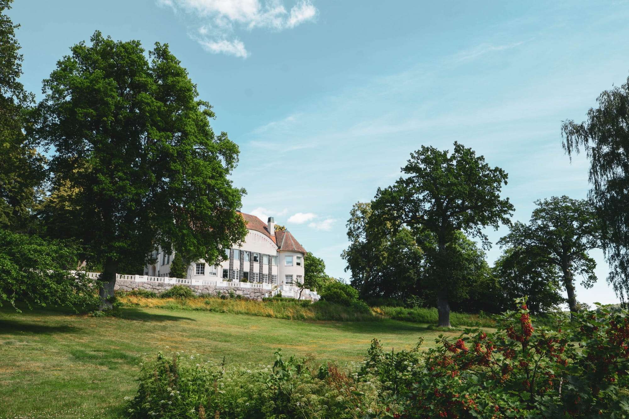 Huset på höjden sett från gräset, med vit balustrad längs terrassen och två stora ekar som ram.