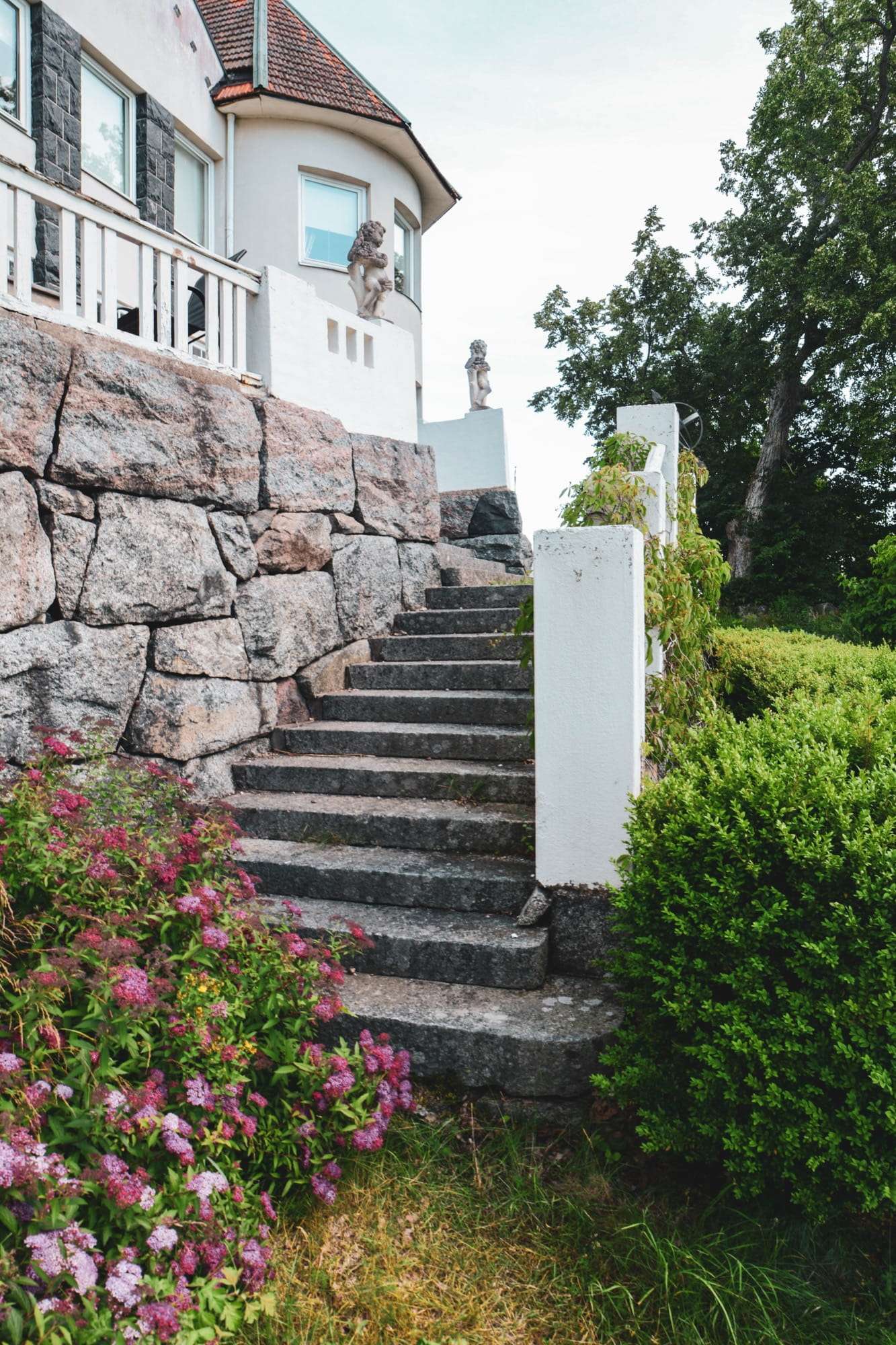 Granittrappan upp från trädgården mot huset, med grov stenmur, vitputsade pelare, putti på balustraden och blommande buskar.