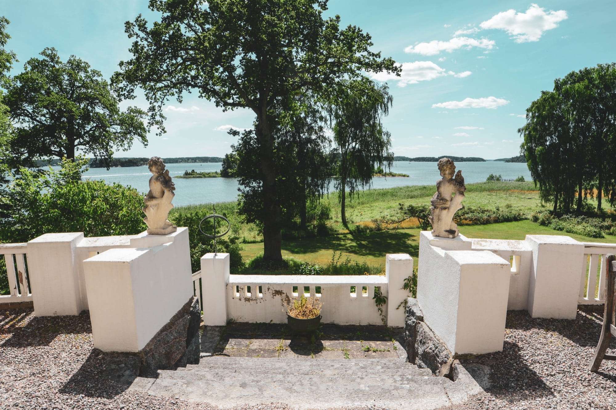 Utsikten från terrasstrappan ned mot sjön. Två putti i sten står på vitputsade balustradpelare, en stor ek mitt i bilden och viken med holmar bakom.