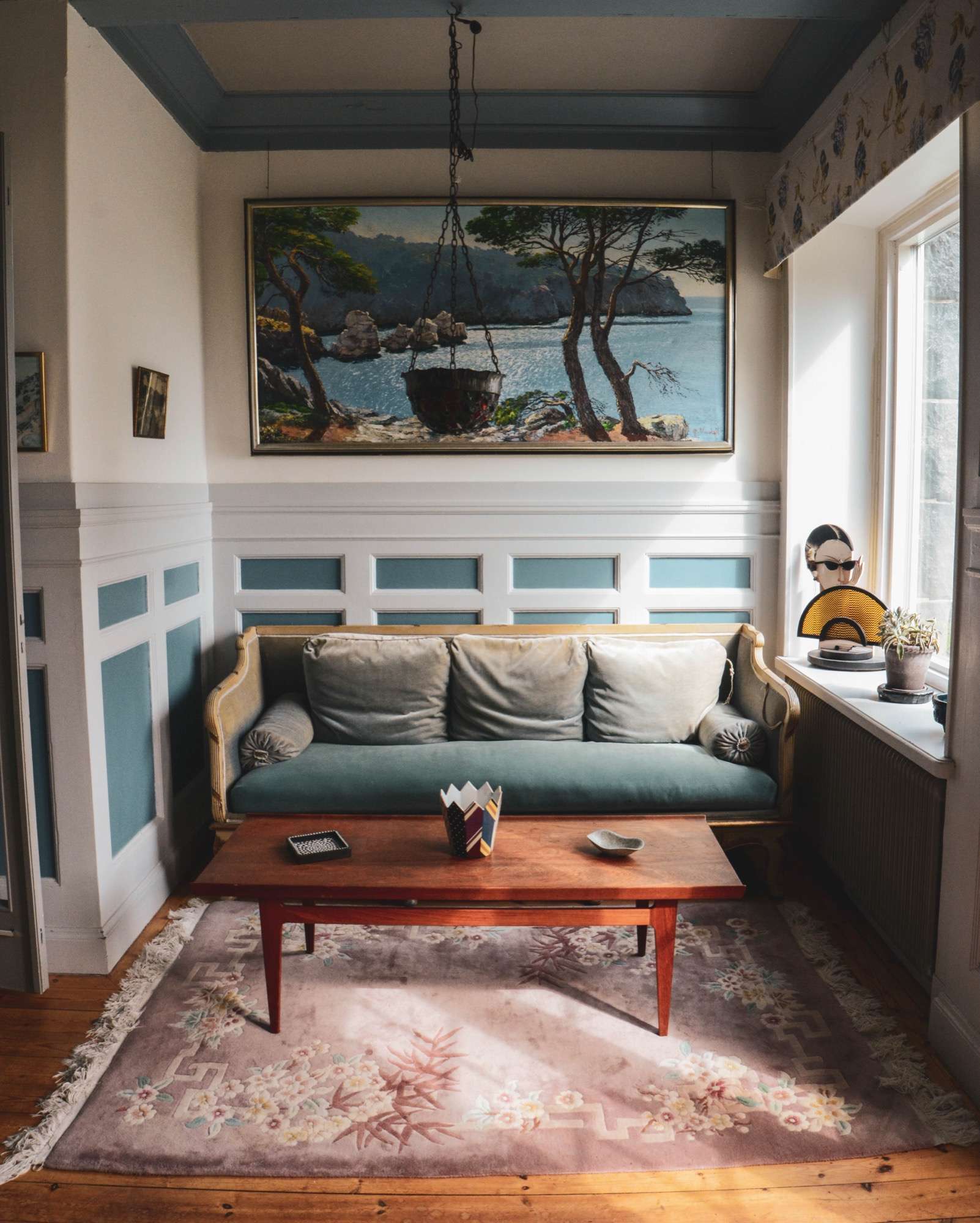 Grön soffa under en stor målning av tallar vid havet, med blå och vit bröstpanel, ett rött soffbord på en ljus matta och en taklampa som hänger över bordet.